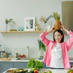 Asian woman in pink overshirt holding a pineapple in a modern kitchen with fresh produce and cheerful ambiance.
